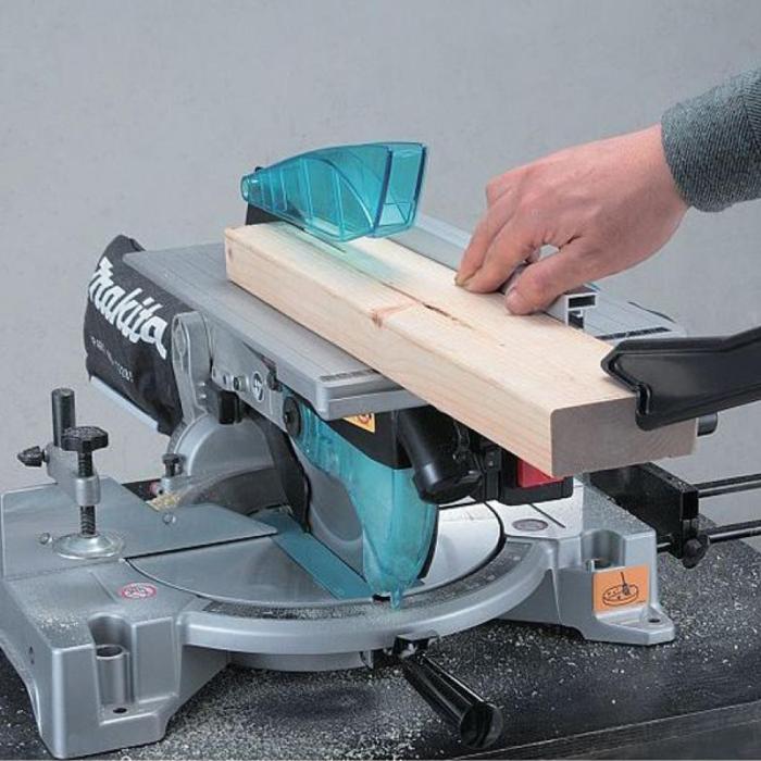 A Makita portable table saw in gray and teal cutting a piece of light wood, with a person's hand guiding the wood feed.