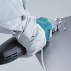A close-up of a person wearing white protective gloves and a white jacket, holding a teal and gray Makita Electric Metal Shear against a sheet of metal.