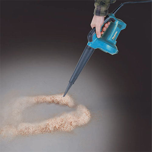A hand holding a blue and black Makita dust blower, creating a swirl of sand on a dark gray surface.