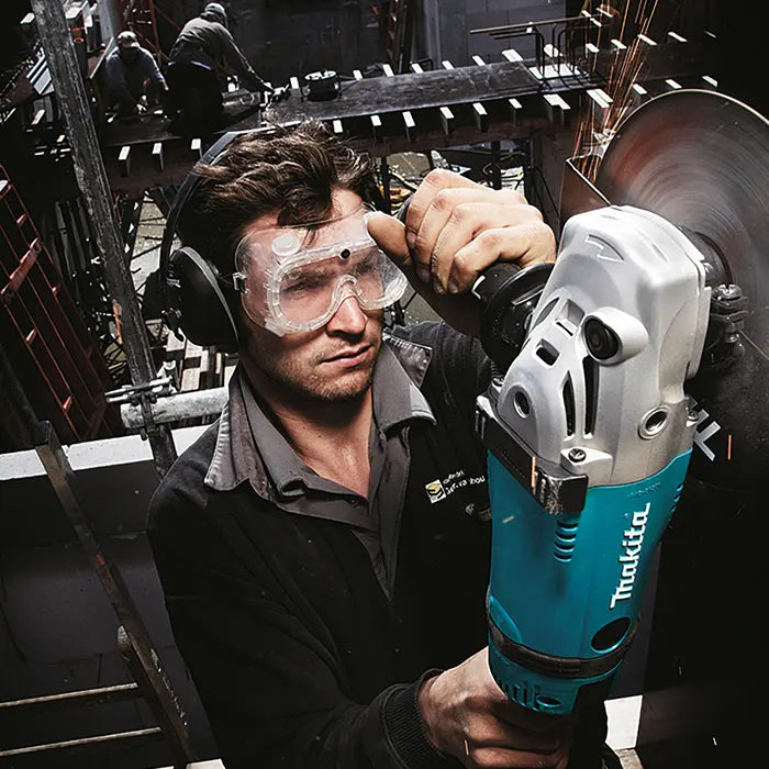 A man wearing safety glasses and ear protection operates a Makita angle grinder on a construction site, with steel beams and other workers visible in the background.