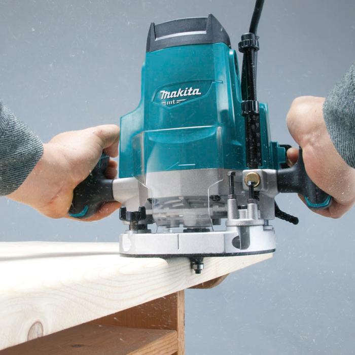 A person using a teal and silver Makita plunge router to trim a light wood board against a gray background.