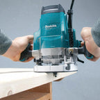 A person using a teal and silver Makita plunge router to trim a light wood board against a gray background.