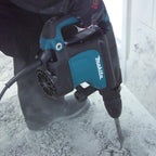 A Makita rotary hammer drill being used by a person to drill into a concrete surface, with dust and debris visible around the drill bit.