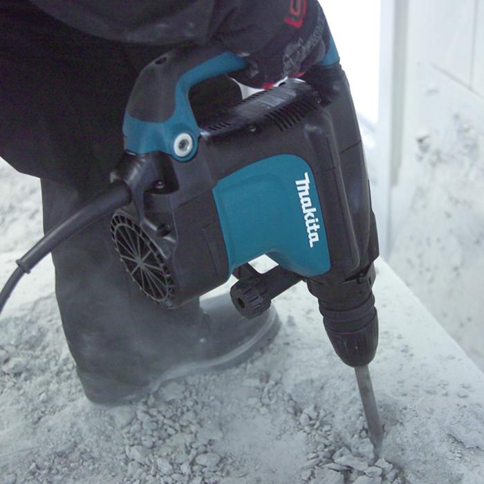 A Makita rotary hammer drill being used by a person to drill into a concrete surface, with dust and debris visible around the drill bit.