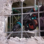 A Makita Concrete Jack Hammer with a metal chisel bit, being operated by a person wearing a black glove on a construction site with concrete rubble and rebar visible.