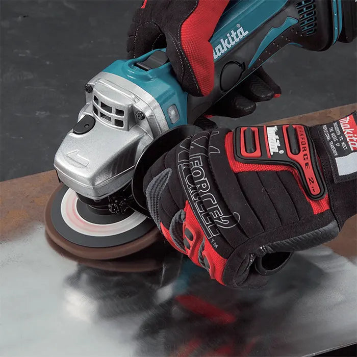 A Makita cordless angle grinder with a sanding disc being held down by a person wearing black and red protective gloves while working on a metal surface.