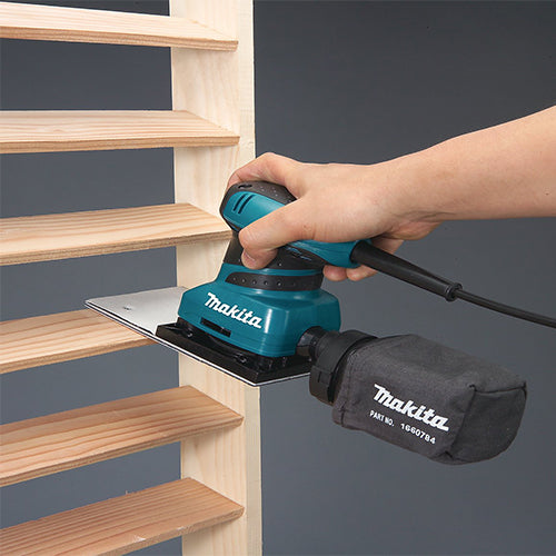 A teal Makita orbital sander being used by a hand to sand a wooden stair tread, with a black Makita dust bag attached. The background is a solid dark gray.