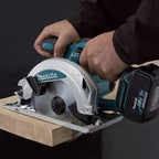 A close-up of a person using a Makita cordless circular saw to cut a piece of wood. The Makita brand is clearly visible on the tool, and the background is a dark, neutral studio setting.