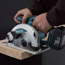 A close-up of a person using a Makita cordless circular saw to cut a piece of wood. The Makita brand is clearly visible on the tool, and the background is a dark, neutral studio setting.