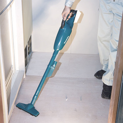 A person using a teal cordless stick vacuum cleaner on a light-colored floor in a doorway or hallway.