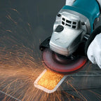A close-up action shot of a teal and silver Makita angle grinder with a black circular blade, cutting through metal and producing bright orange sparks. A white-gloved hand is visible on the right side of the grinder.