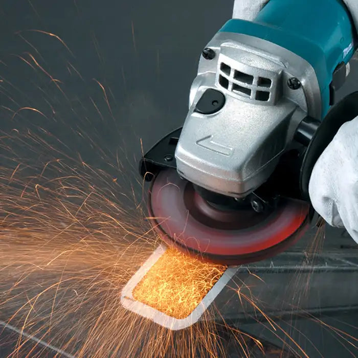 A close-up action shot of a teal and silver Makita angle grinder with a black circular blade, cutting through metal and producing bright orange sparks. A white-gloved hand is visible on the right side of the grinder.