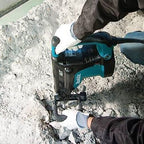 A Makita cordless jackhammer drill being operated by a person wearing white work gloves and a dark jacket, breaking concrete with the tool in an outdoor or industrial setting.