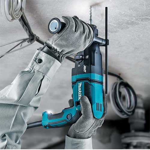 A person wearing gray work gloves uses a teal and black Makita cordless drill hammer on a ceiling in a construction setting.