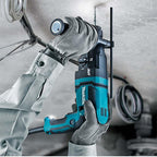 A person wearing gray work gloves uses a teal and black Makita cordless drill hammer on a ceiling in a construction setting.