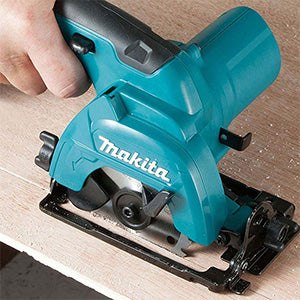 A teal and black Makita circular saw with a guide rail, shown in use with a person's hand on a piece of wood.