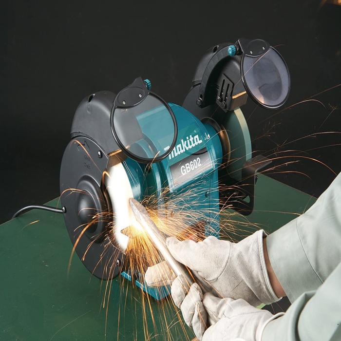 A Makita bench grinder in teal and black, shown in use with sparks flying as a person wearing white gloves holds a metal rod against the cutting wheel. The grinder has protective guards and a visible Makita logo.