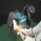 A Makita bench grinder in teal and black, shown in use with sparks flying as a person wearing white gloves holds a metal rod against the cutting wheel. The grinder has protective guards and a visible Makita logo.