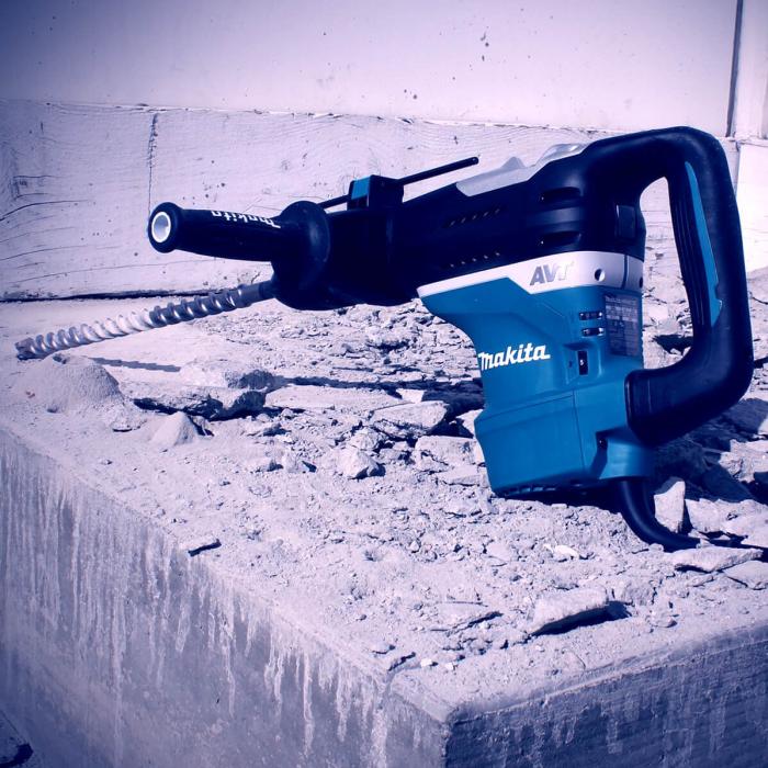 A Makita rotary hammer drill with a spiral coring bit, positioned on a concrete ledge with rubble in the foreground.
