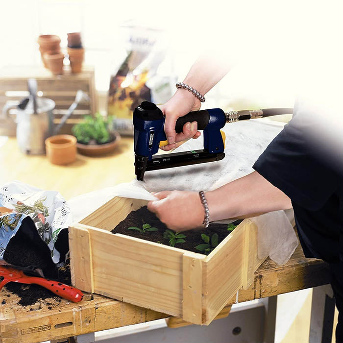 A person using a blue and yellow pneumatic nail gun to fasten a white liner into a wooden raised garden bed on a workbench, with gardening tools and pots in the background.