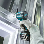 A person wearing a light gray work glove holds a Makita impact driver against a galvanized steel wall or ceiling during installation.