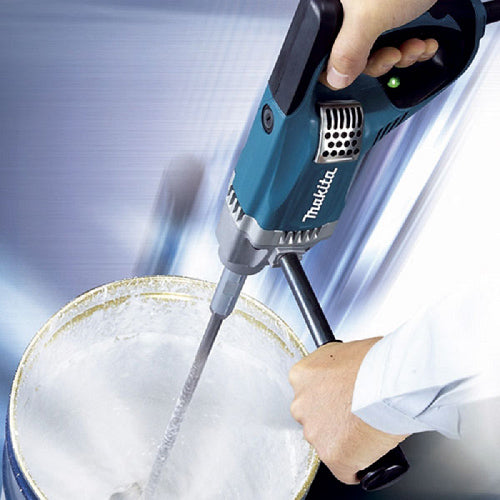 A Makita cordless electric paint mixer held by a person and used to mix a white liquid inside a bucket, shown against a blue and white blurred background.