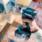 A person's hand holding a Makita cordless impact driver while installing metal wall studs into a wooden wall frame.
