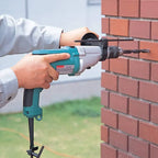 A person using a teal and gray cordless Makita rotary hammer drill to drill into a red brick wall outdoors.