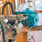 A miter saw with a teal circular blade guard and silver metal components, shown in a workshop setting with a person's tattooed arm guiding a wood board through the blade.