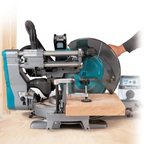 Makita miter saw with a teal and gray body, shown cutting a piece of wood. A human arm is visible pushing the saw, and a dust collector is connected.