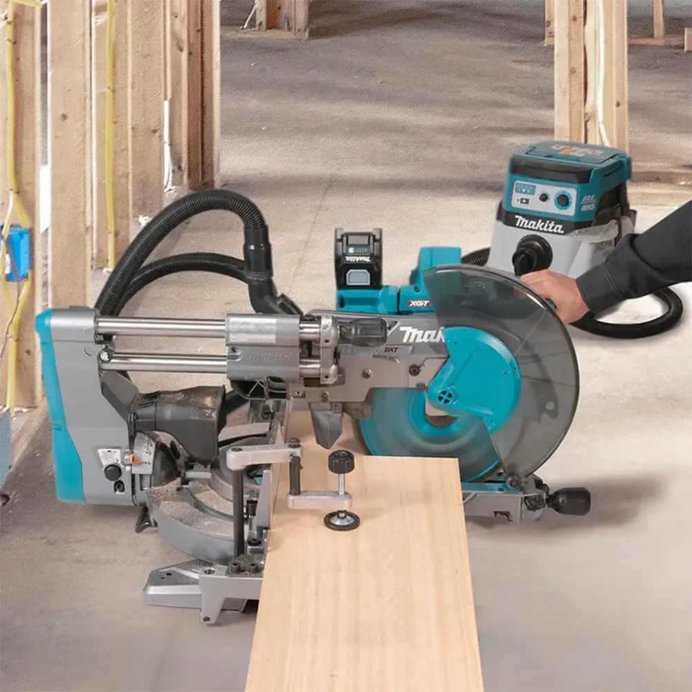 Makita miter saw cutting a piece of light wood, with a Makita shop vacuum in the background on a concrete floor inside a construction site.