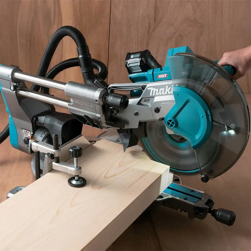 A Makita XDT 10-in-1 miter saw cutting a piece of light wood, with a person's hand visible on the handle. The saw features a teal and silver body with a clear dust cover, set against a wooden background.