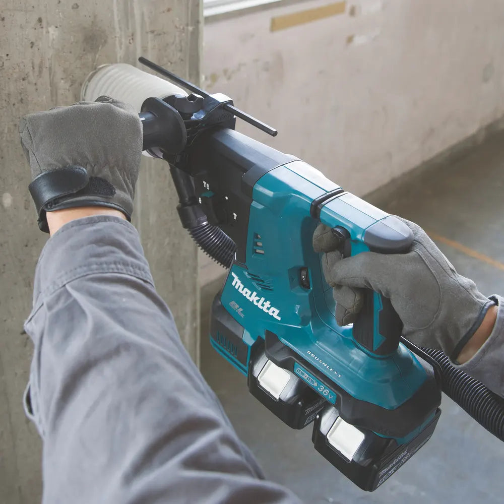 A person wearing gray work gloves operates a teal and black Makita cordless rotary hammer against a concrete wall in an industrial setting.