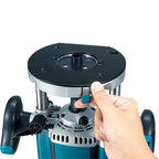 A close-up of a Makita Wood Router Machine with a black and silver base plate, teal body, and black handles, with a hand adjusting a small teal switch on the front.