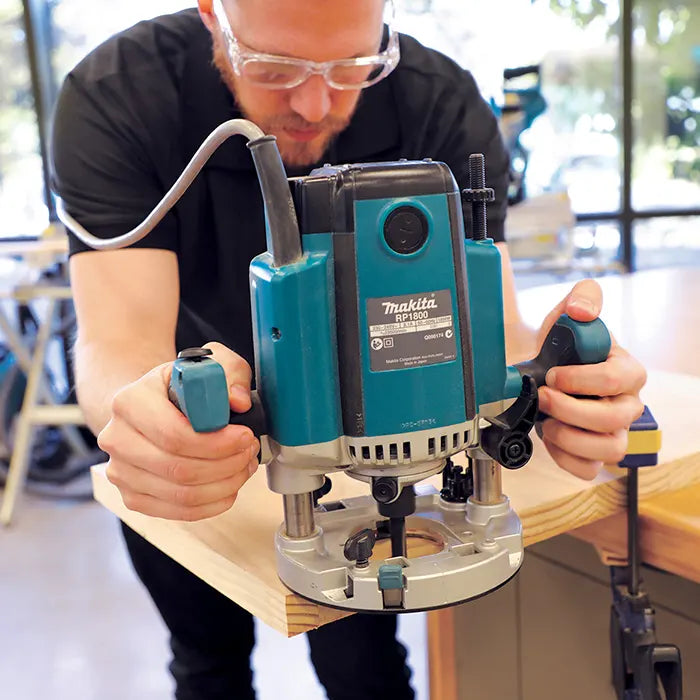 A man wearing safety glasses uses a teal and silver Makita Wood Router Machine to carve a wooden board in a workshop setting.