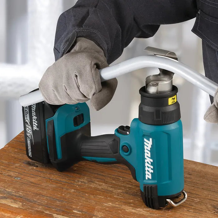 A person wearing gray work gloves operates a teal and black Makita cordless hot gun on a wooden workbench, with a metal pipe fed into the chuck.
