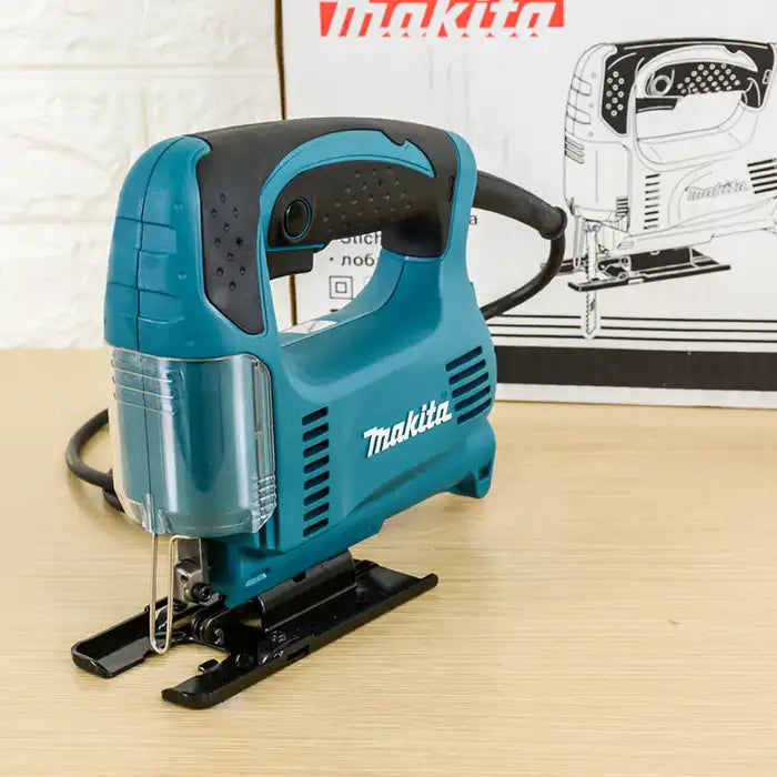 A Makita jigsaw saw with a teal and black body, shown on a light wood surface with its product box in the background.