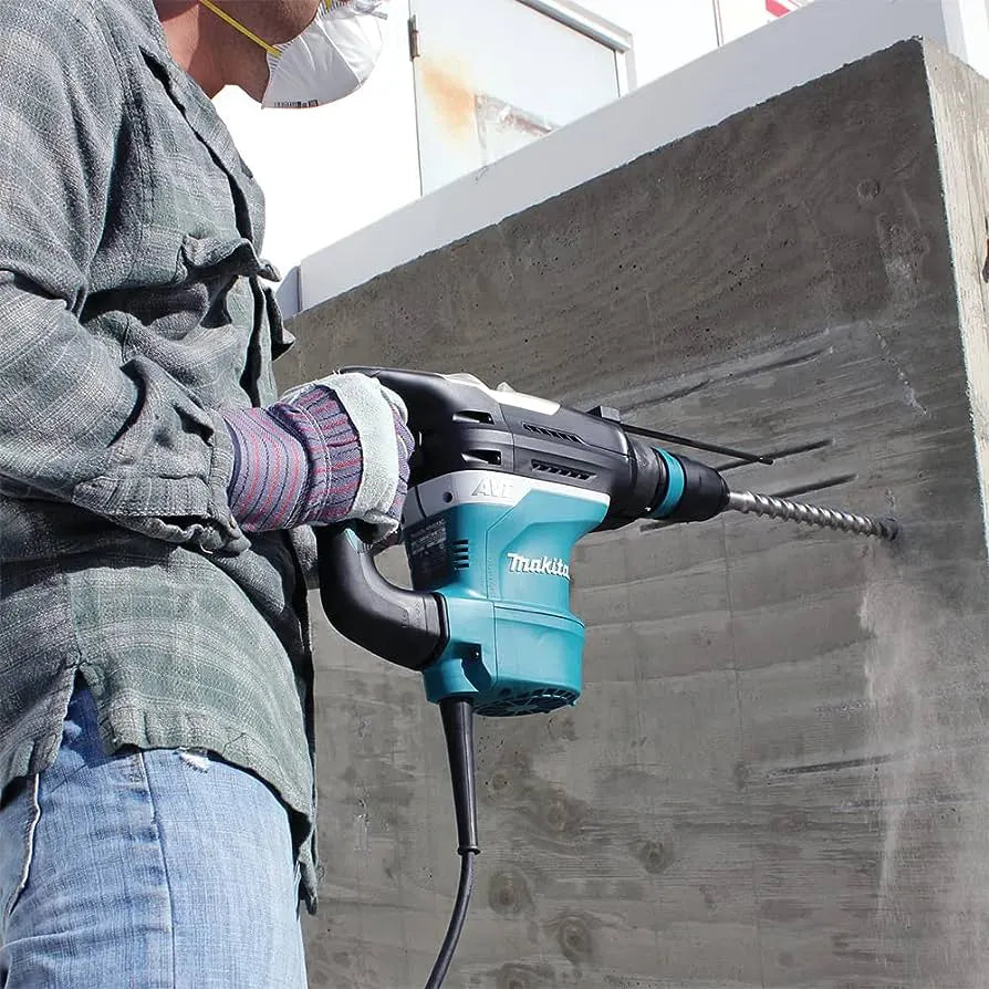 A person wearing protective gear uses a teal and black Makita rotary hammer drill with a spiral hole saw attachment to drill into a large concrete wall.
