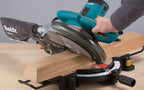 A teal Makita miter saw in use, cutting a light wood board on a workbench with a person's hand guiding the blade.